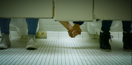 The open bottom of two bathroom stalls with a person in each stall reaching underneath to hold the hand of the person in the stall next to them.